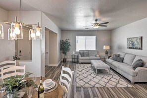 A modern living room with a grey sofa, a dining table with chairs, a ceiling fan, and a large window with blinds. - Idyllic Kingman Retreat With Yard - Near Hiking (Kingman)