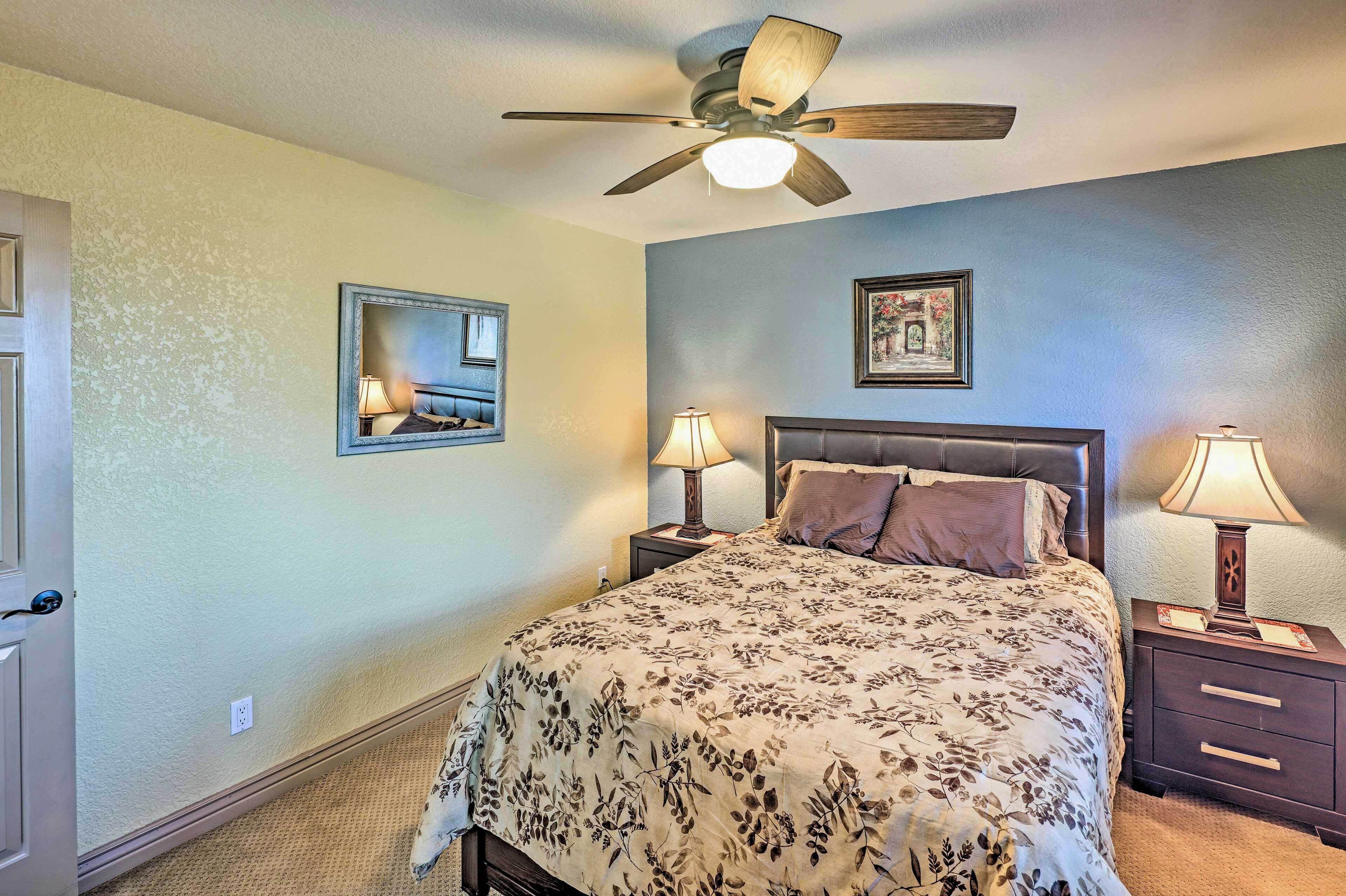 A bedroom with a bed, two bedside tables with lamps, a ceiling fan, a door, and a wall-mounted mirror.