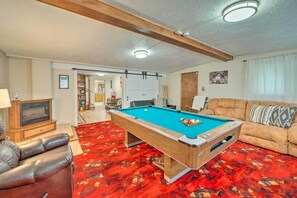A pool table in a living room with a red carpet, a leather sofa, and a wooden ceiling beam.