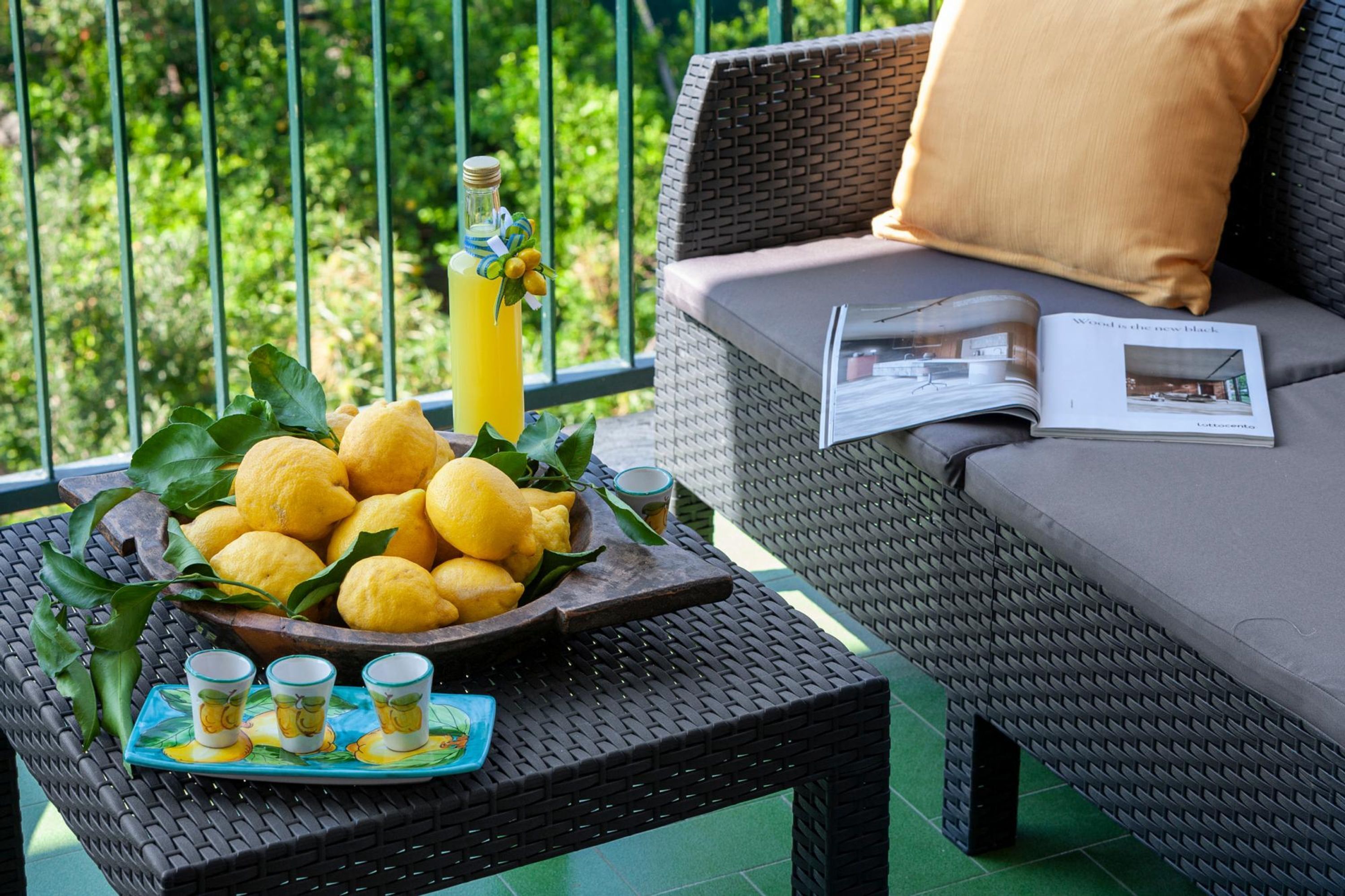 A wicker table with a bowl of lemons, a bottle of lemonade, and shot glasses on a balcony with a green railing and a sofa with a magazine.