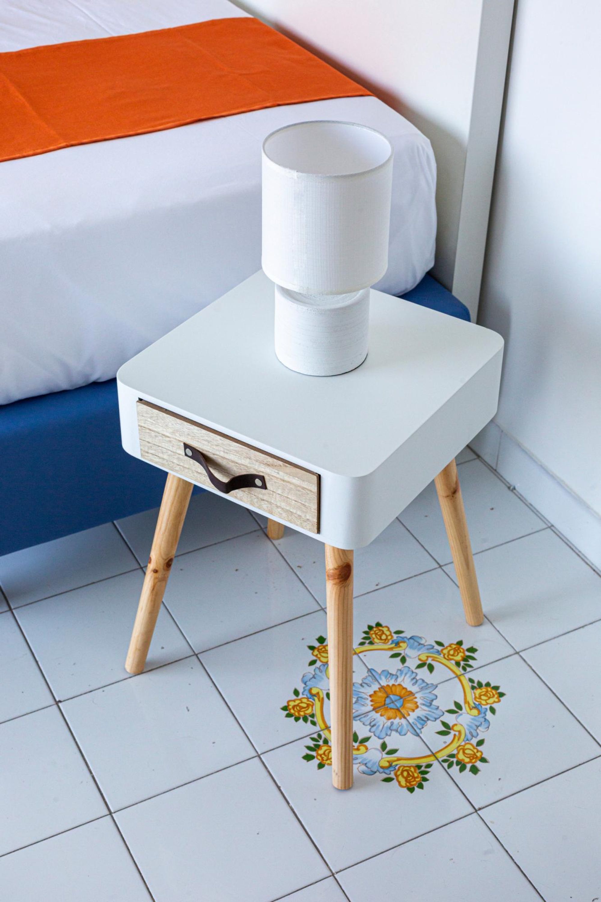 Una pequeña mesita de noche blanca con un cajón, una lámpara blanca y un azulejo decorativo con motivos florales.
