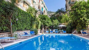A swimming pool with blue lounge chairs and umbrellas, surrounded by greenery and buildings. - Casa Maridea in Sorrento (Sorrento)
