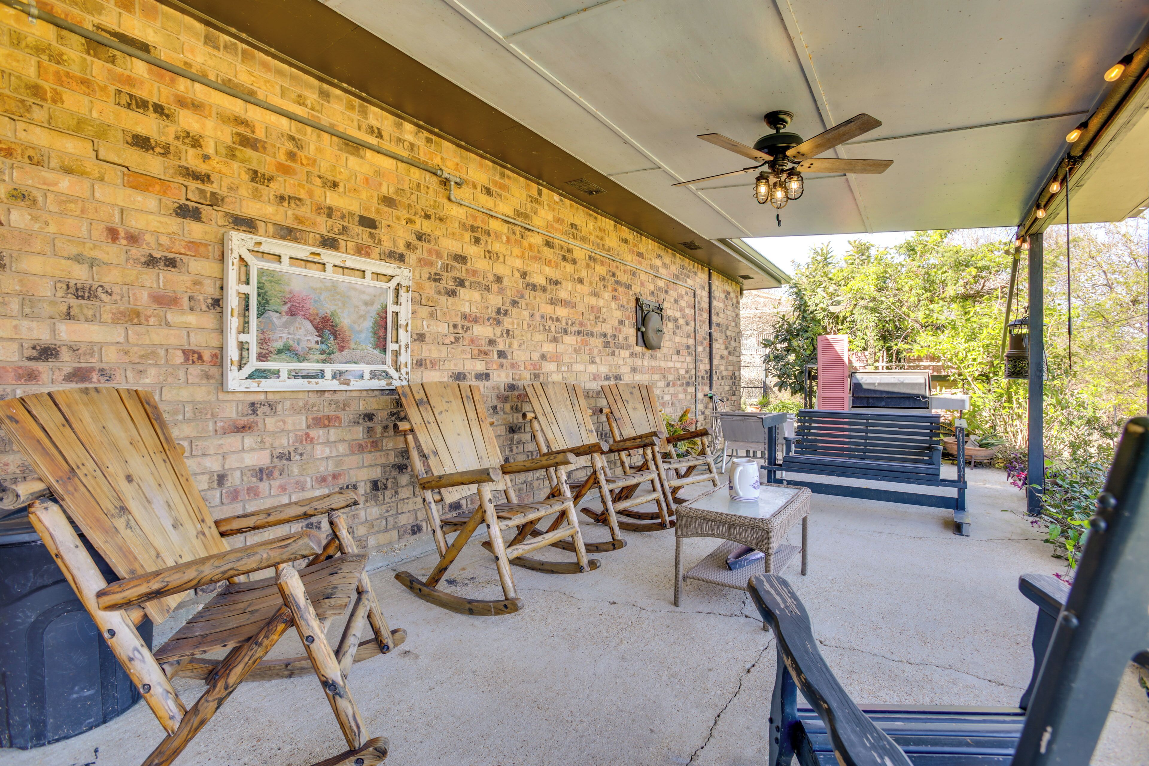 A covered patio area with brick walls, wooden rocking chairs, a table, and a ceiling fan.