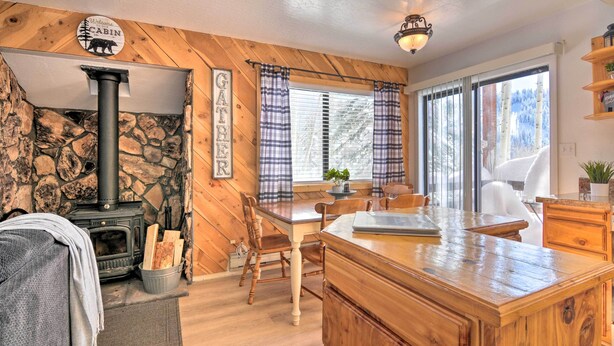 Ein Innenraum einer Hütte mit einer Steinsandsteinwand, einem Holzofen, einem Essbereich mit einem Holztisch und Stühlen sowie einem großen Fenster mit Vorhängen.