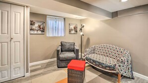 A living room with a grey armchair, a covered sofa, a red ottoman, and a floor lamp. There are two paintings on the wall and a door on the left. - Garden-level Millburn Apt - Walk to Transit! (Millburn)
