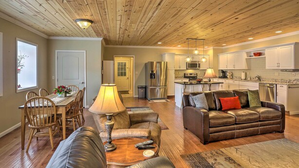 A spacious living room with a wooden ceiling, a dining area with a table and chairs, a leather sofa, and a kitchen with white cabinets and stainless steel appliances.