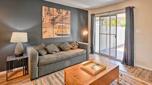 A living room with a grey sofa, a wooden coffee table, a lamp, and a large window with curtains. - Scottsdale Townhome w/ Pool Access, Near Old Town! (Scottsdale)