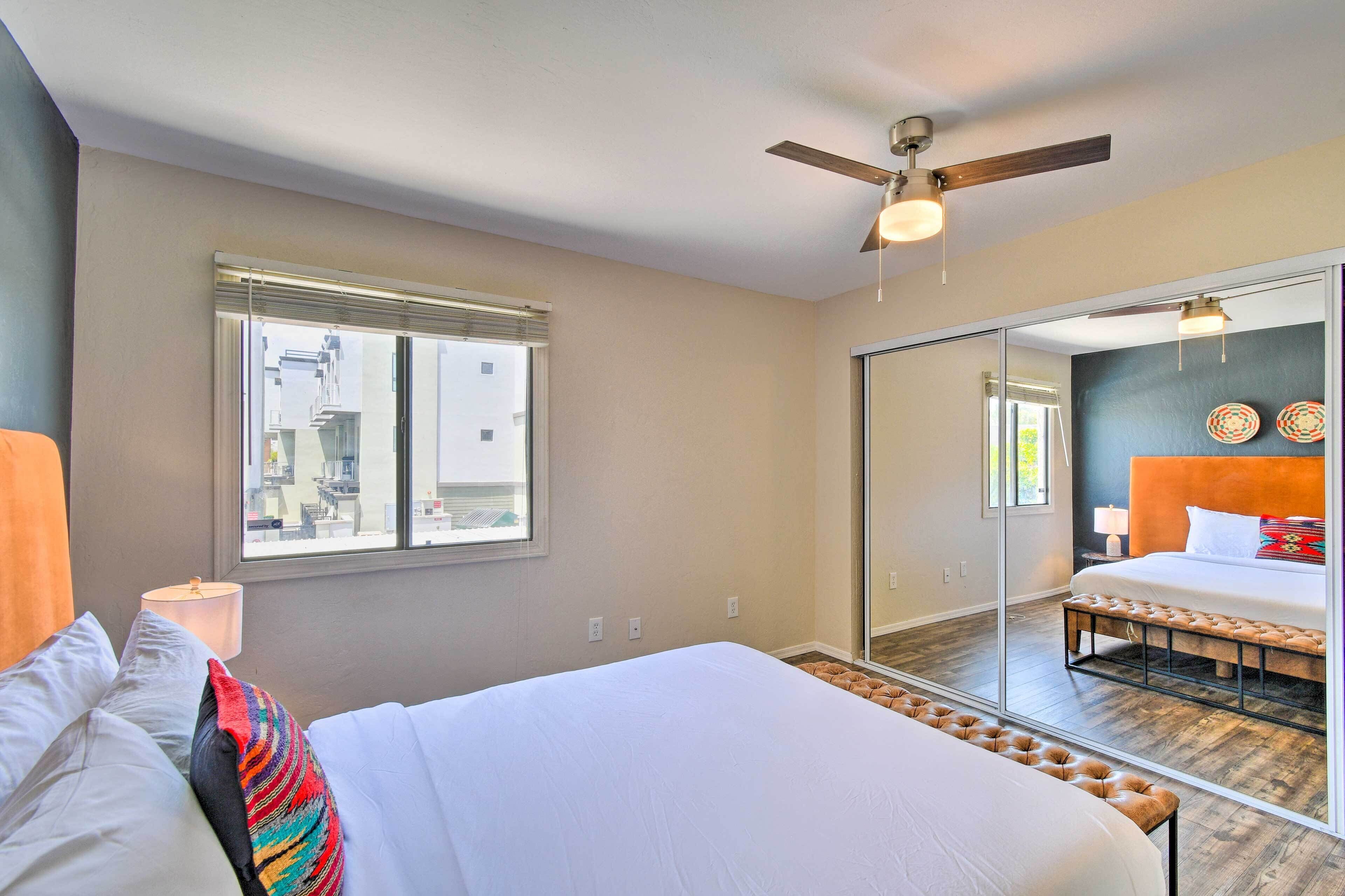 Un dormitorio con una cama, ventilador de techo, ventana y espejo que refleja la habitación.