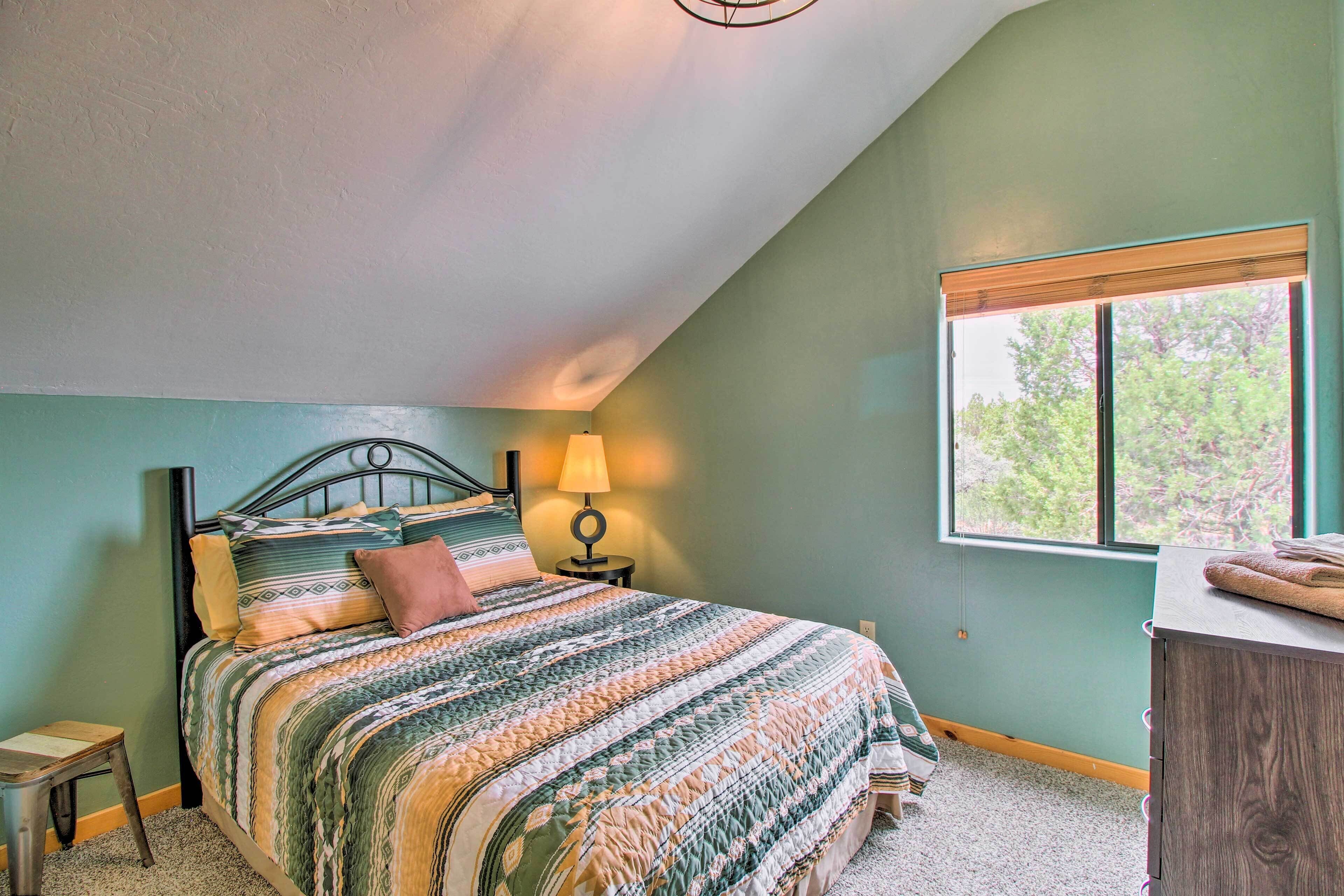 A bedroom with a sloped ceiling, a bed with a patterned quilt, a bedside lamp, a window with a view of trees, and a wooden dresser.