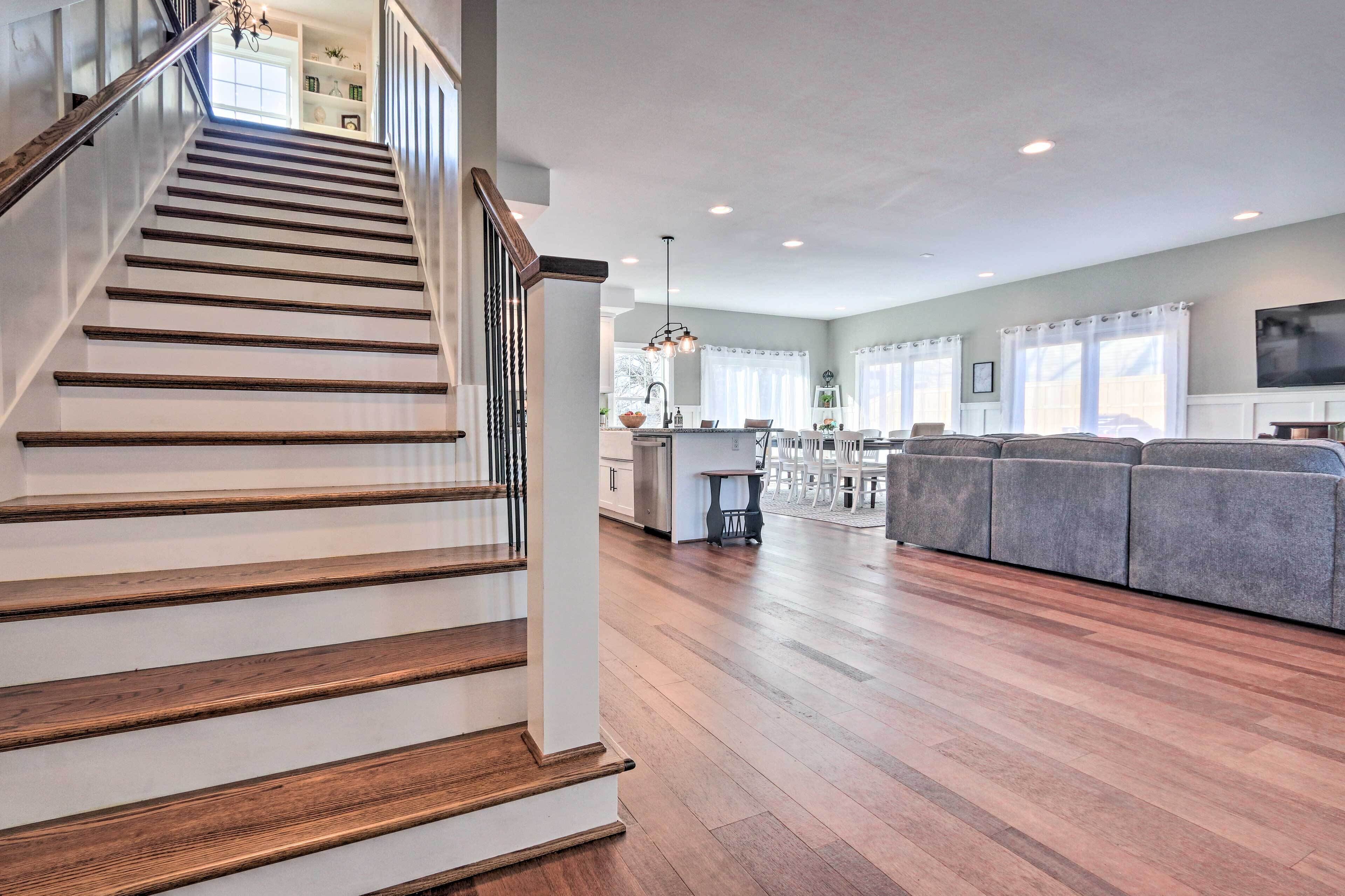 A spacious living area with a wooden staircase, a gray sofa, and a kitchen in the background.