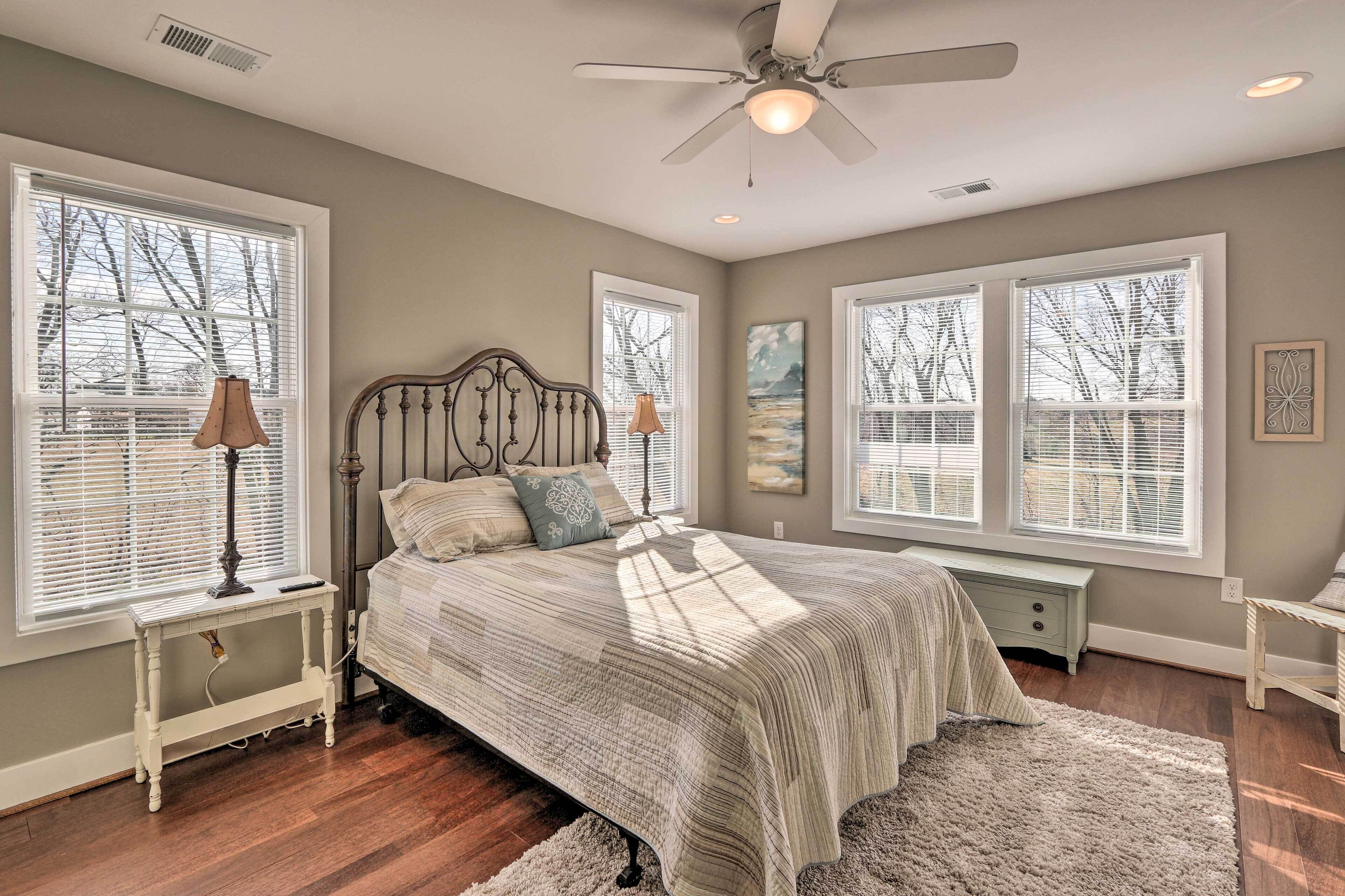 A bedroom with a bed, two bedside tables, a ceiling fan, and large windows with blinds.