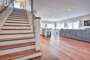 Uma sala espaçosa com escada de madeira, um sofá cinzento e uma cozinha ao fundo.