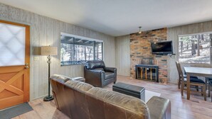 Un salon chaleureux avec un canapé en cuir, une cheminée, un téléviseur fixé au mur en briques et de grandes fenêtres donnant sur la forêt.