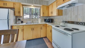 Une cuisine compacte avec des meubles en bois, un réfrigérateur blanc, une cuisinière et une fenêtre donnant sur la forêt.