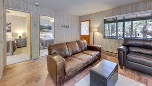 Un salon avec un canapé en cuir marron, un pouf assorti et un plancher en bois. Une porte donne accès à une chambre à coucher où se trouvent un lit et une table de chevet.