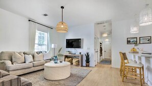 Een moderne woonkamer met een bank, een poef en houten stoelen. Een televisie is aan de muur bevestigd, en er staat een klein tafeltje met decoratieve objecten.