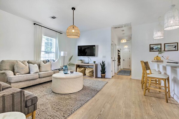 Un salon moderne avec un canapé, un pouf et des chaises en bois. Un téléviseur est fixé au mur, et une petite table est agrémentée d’objets décoratifs.