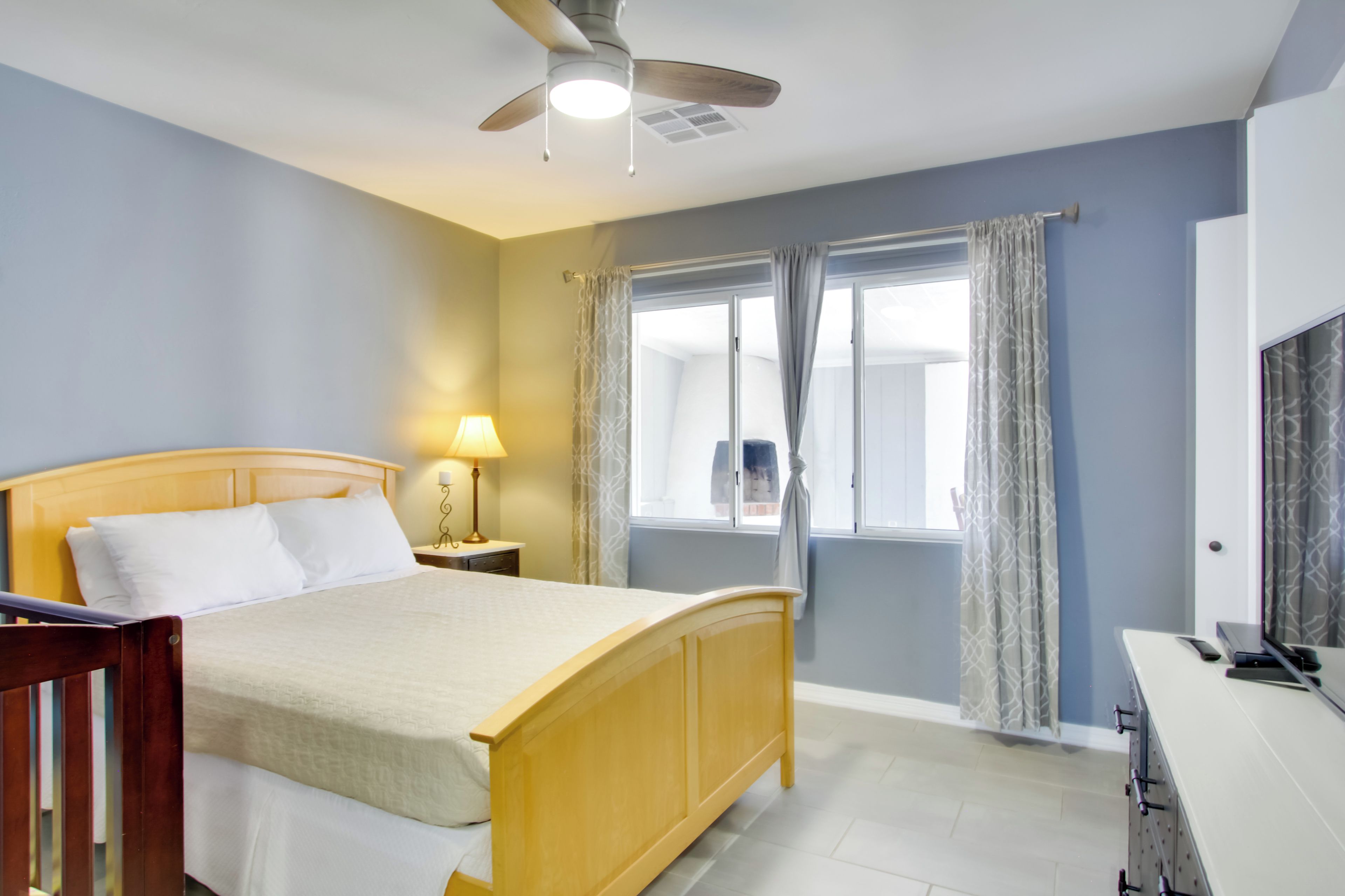 A bedroom with a wooden bed, a nightstand, a lamp, a ceiling fan, a window with curtains, and a TV on a dresser.