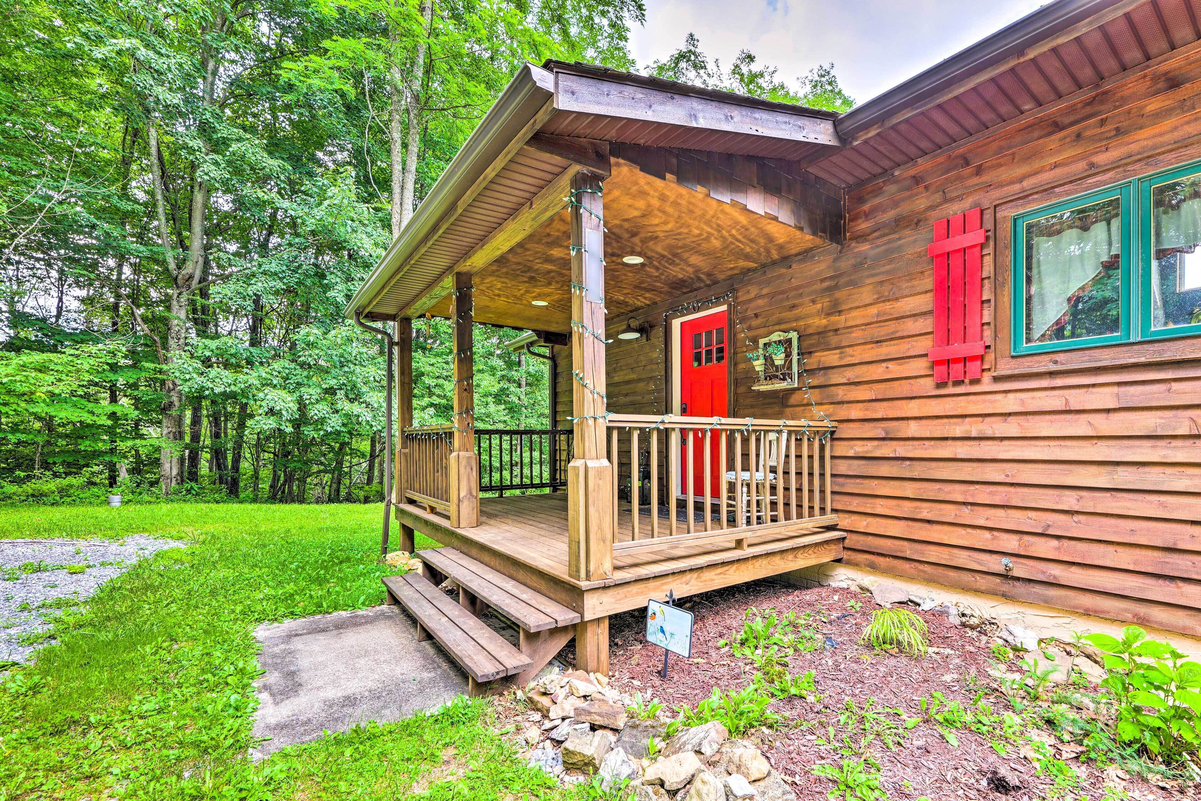 A wooden cabin with a porch, a red door, and a view of a forested area.