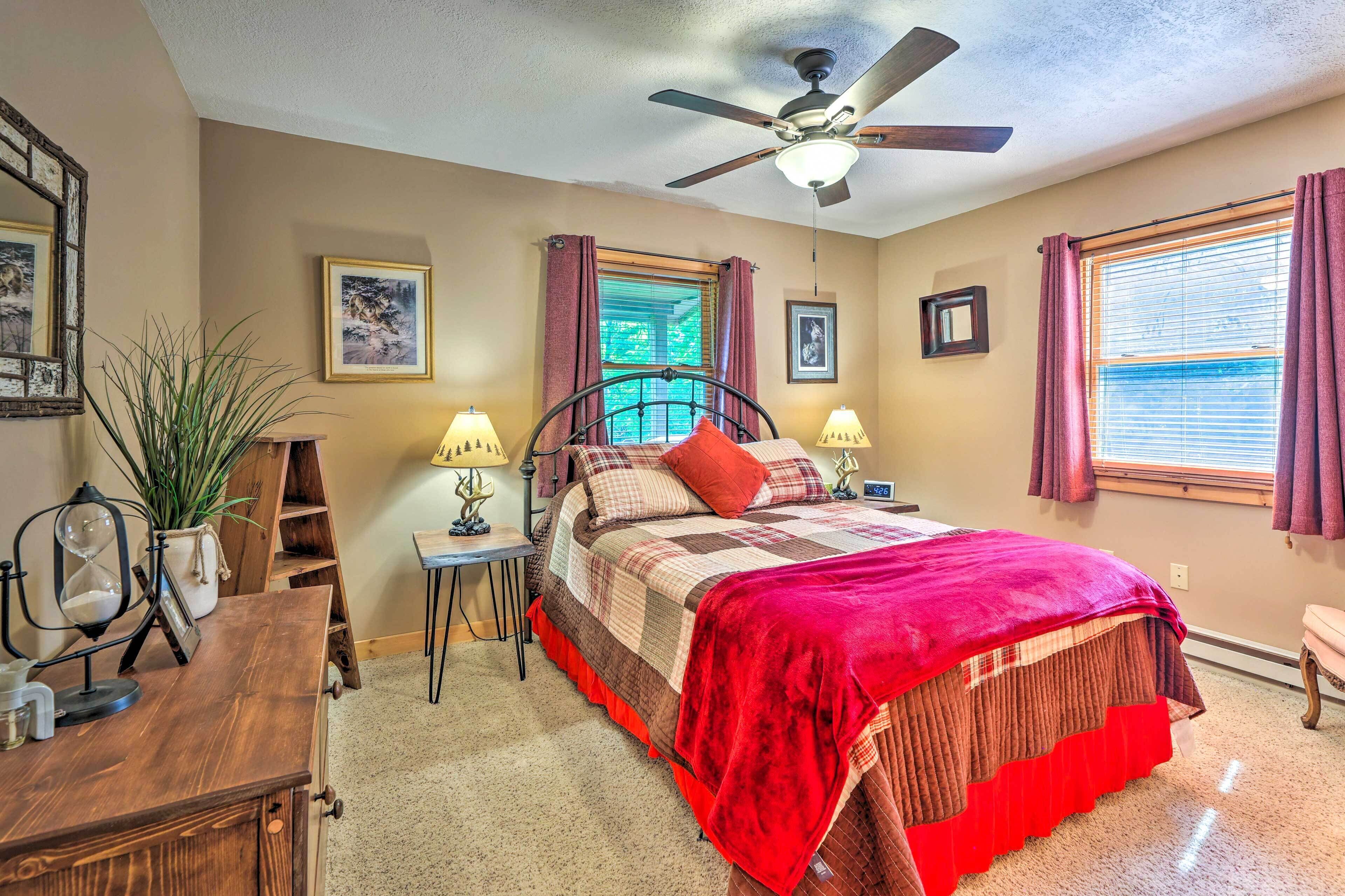 A bedroom with a bed, ceiling fan, two windows with curtains, a desk with a lamp, and a potted plant.