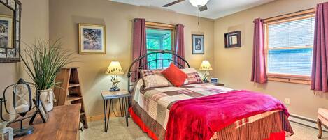 A bedroom with a bed, ceiling fan, two windows with curtains, a desk with a lamp, and a potted plant.