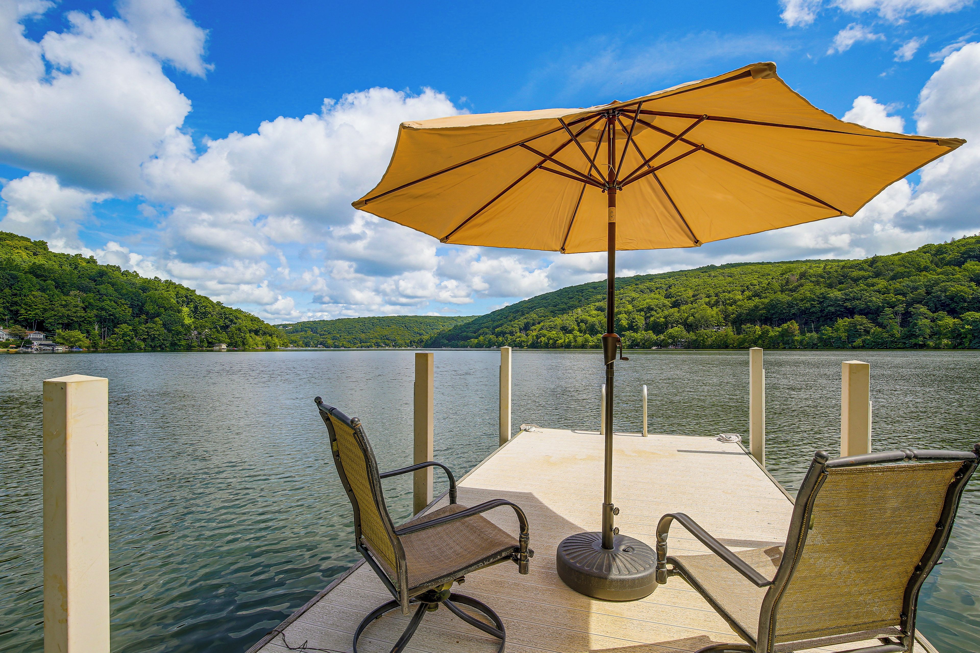 A dock with two chairs and a yellow umbrella, overlooking a calm lake with a forested hill in the background.