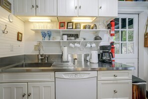 Cozinha com armários brancos, bancada de aço inoxidável, máquina de café e janela com vista para vegetação.