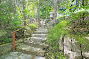 Escadaria de pedra rodeada por vegetação exuberante, levando a um edifício com parede de pedra e corrimão de madeira.