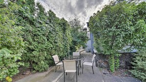 A patio with a glass-top table and four chairs, surrounded by dense greenery and a stone wall. - Bright & Airy Liberty Lake Abode w/ Backyard! (Liberty Lake)