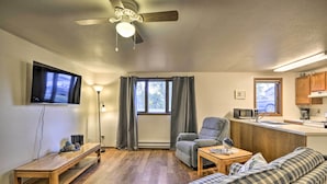 A compact living space with a wooden bench, a sofa, a chair, a television, a ceiling fan, a window with curtains, and a kitchenette area. - Charming Columbus Apt Along Yellowstone River (Columbus)