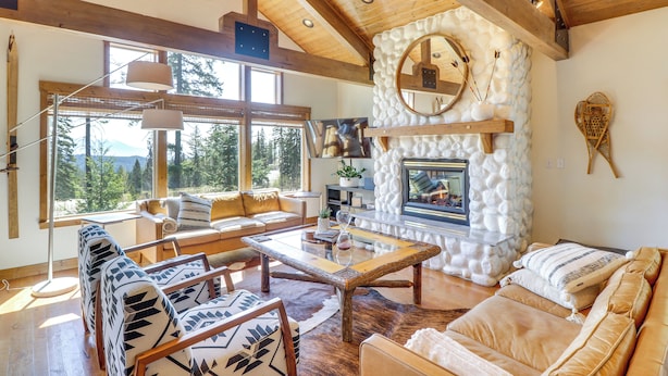 A spacious living room with a stone fireplace, wooden beams, and large windows offering a view of the outdoors.