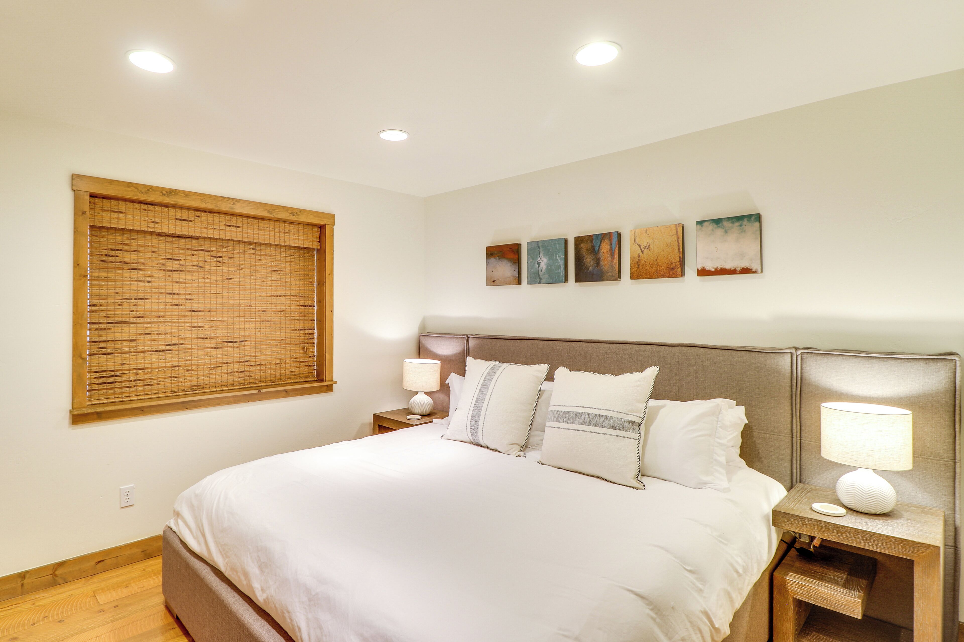 Une chambre bien rangée, avec un lit, des tables de chevet, une fenêtre avec des stores et des œuvres d’art accrochées au mur.