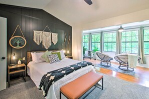 Une chambre moderne avec un grand lit, des tables de chevet, un banc, une chaise et une vue sur la verdure grâce à de grandes fenêtres.