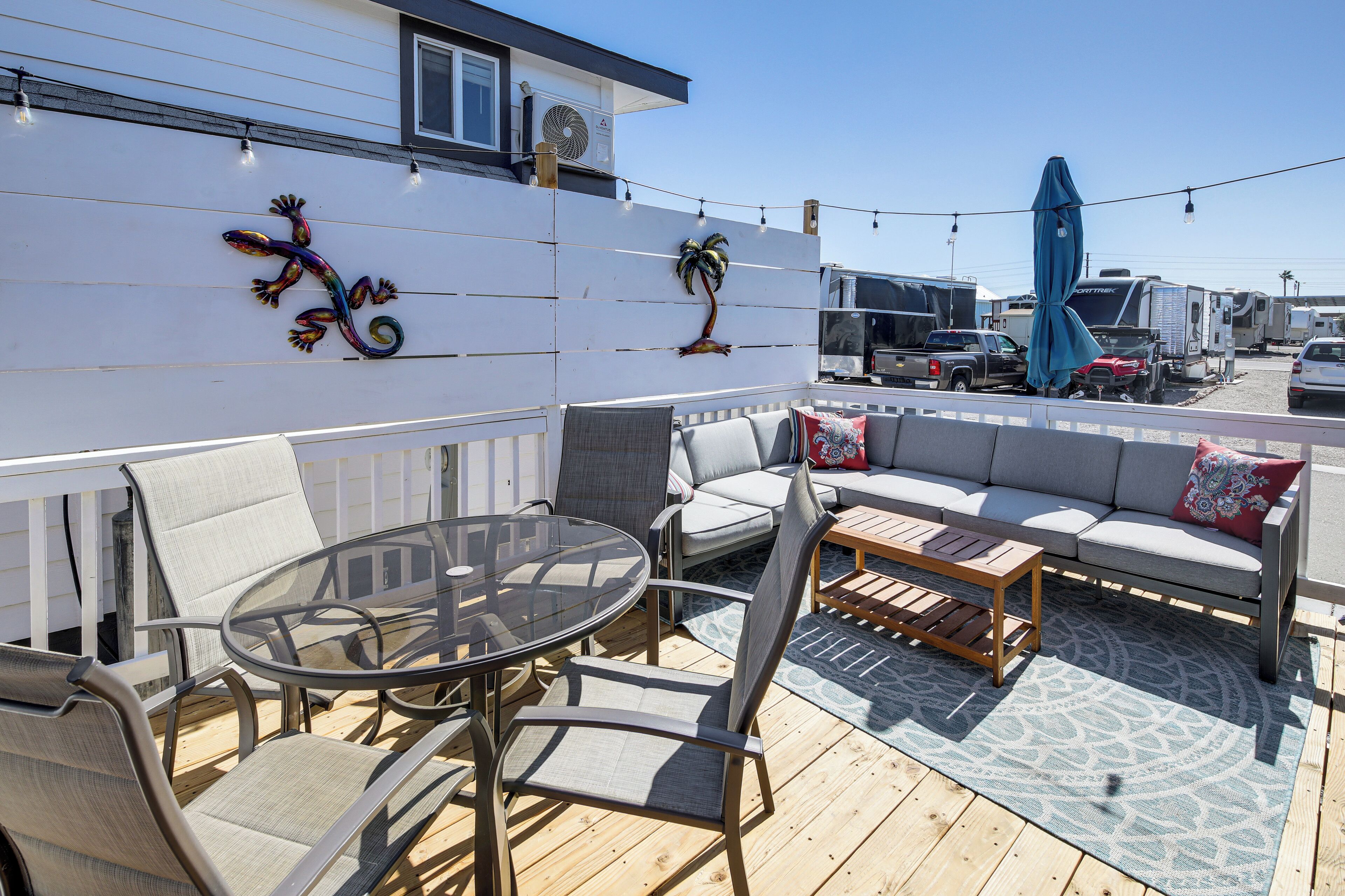 Een terras buiten met een glazen tafel, beklede zitplaatsen en decoratieve muurkunst.