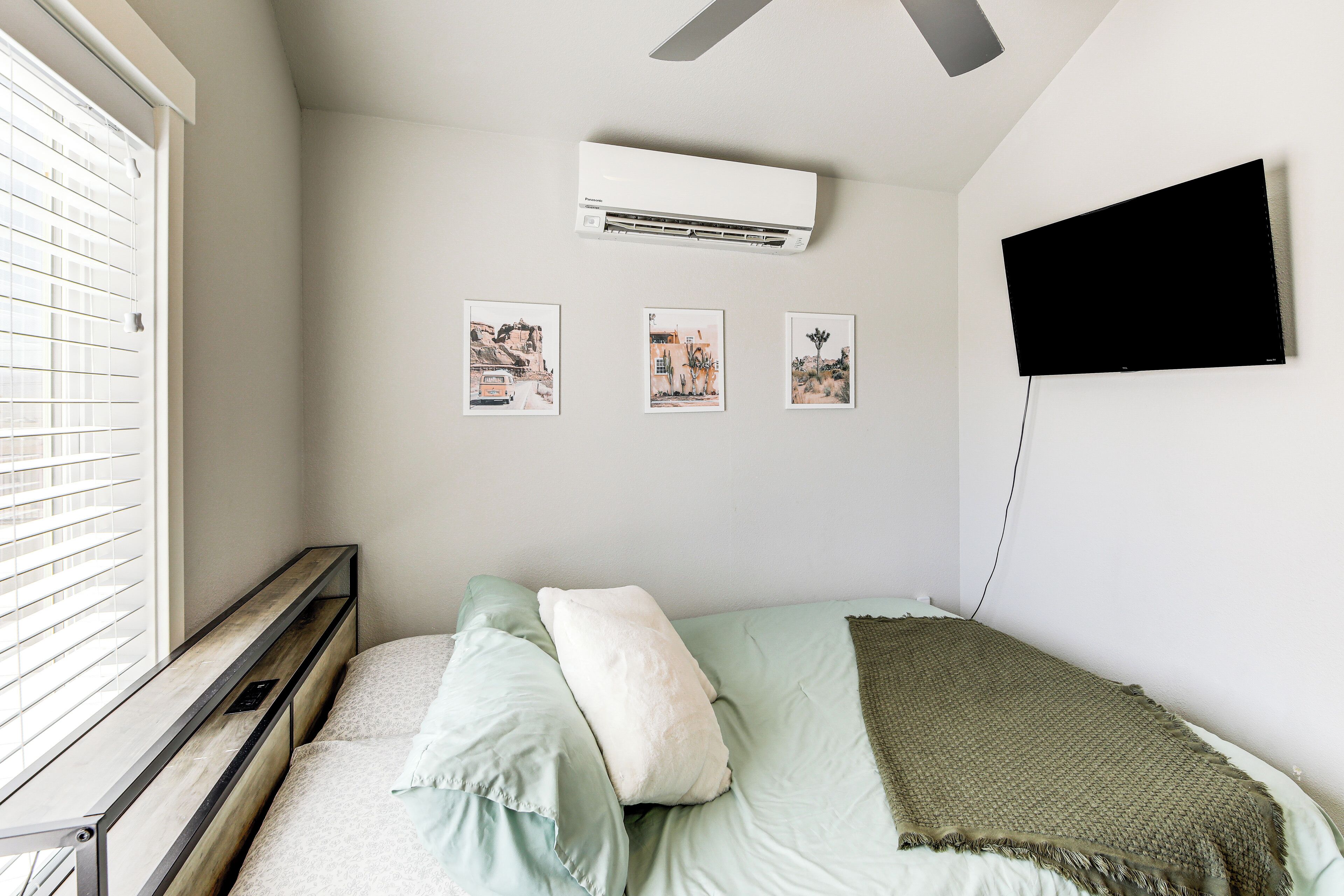 A bedroom with a bed, a window with blinds, an air conditioner, and three framed pictures on the wall.
