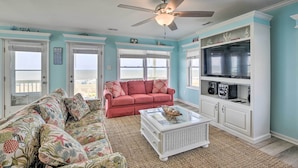 A living room with a red sofa, a patterned couch, a glass coffee table, and a ceiling fan. - Large-group Getaway - Beachfront Home w/ Pool! (Supply)