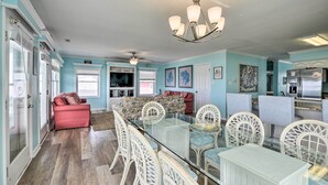 A spacious living room with a glass dining table, white wicker chairs, a red sofa, and a ceiling fan. - Large-group Getaway - Beachfront Home w/ Pool! (Supply)