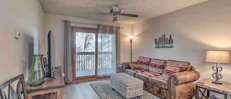 A cozy living room with a sofa, ottoman, and a coffee table. There is a ceiling fan, a lamp, and a decorative wall piece.