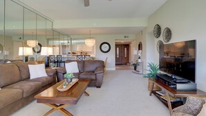 A spacious living room with a large sofa, a coffee table, and a TV. There is a dining area in the background with a chandelier. - Resort Amenities: Rancho Mirage Country Club Home (Rancho Mirage)