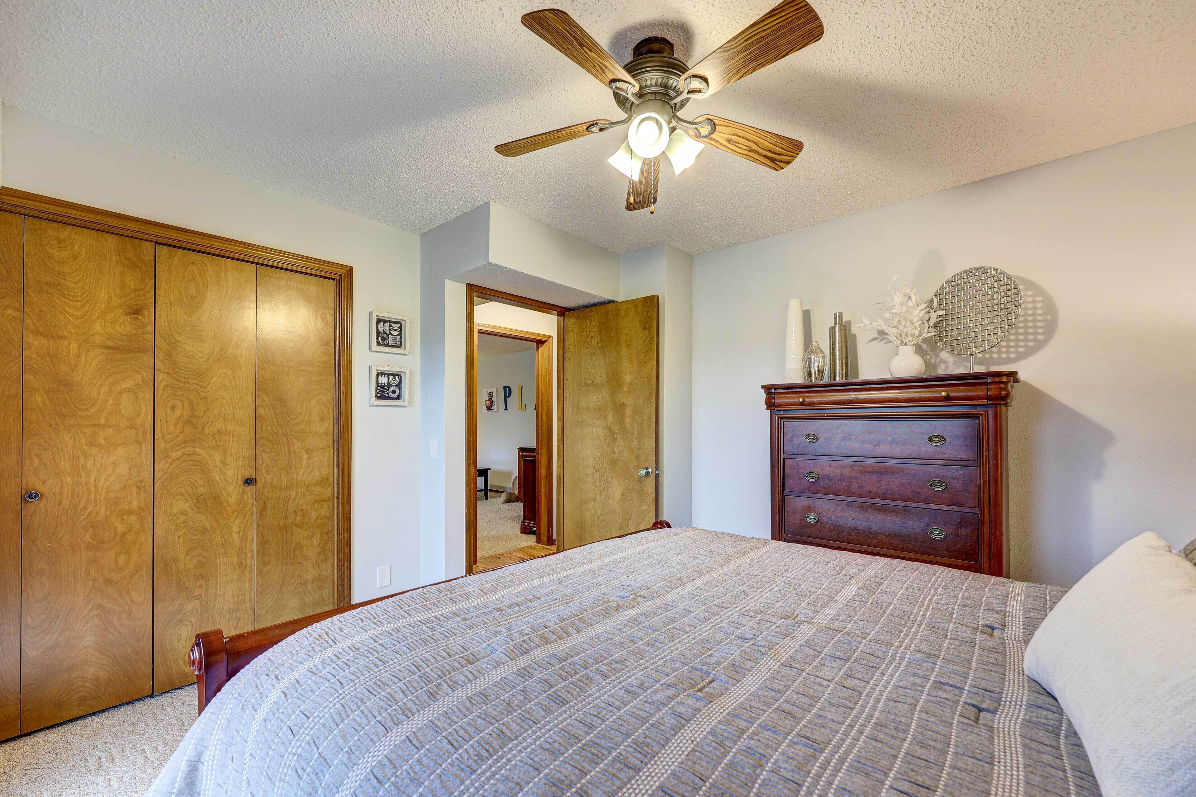 A bedroom with a bed, wooden wardrobe, dresser, ceiling fan, and a door leading to another room.