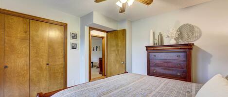 A bedroom with a bed, wooden wardrobe, dresser, ceiling fan, and a door leading to another room.