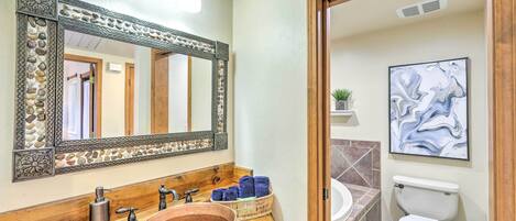 A bathroom with a copper sink, wooden vanity, decorative mirror, and a modern abstract painting.