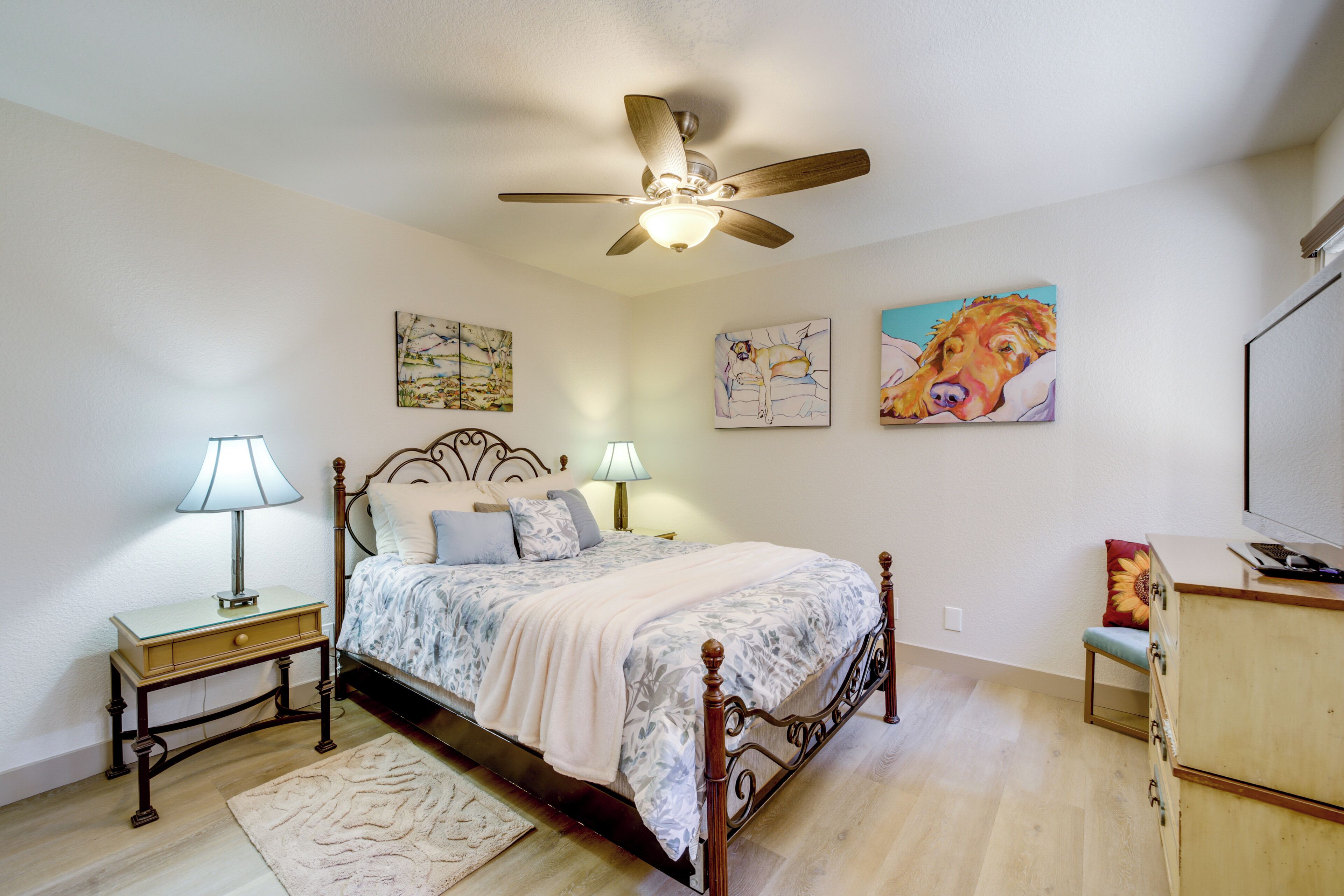A bedroom with a bed, bedside tables, a ceiling fan, artwork on the walls, and a dresser.