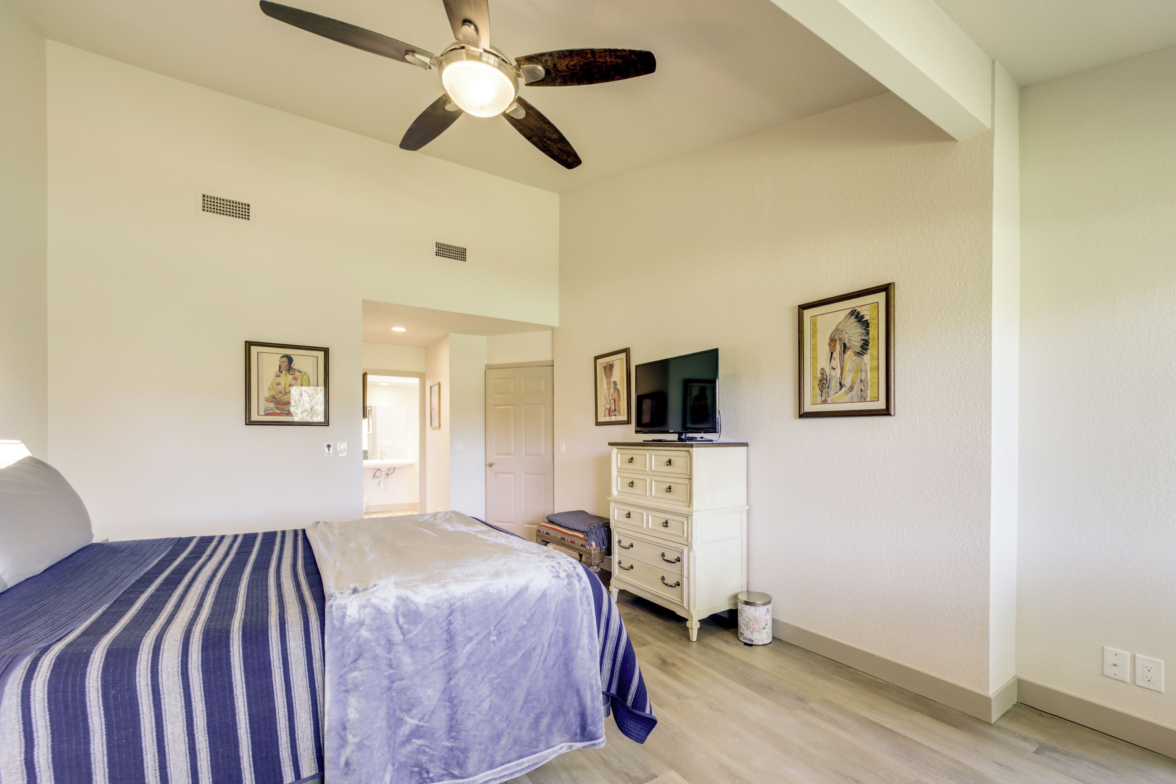 A bedroom with a bed, dresser, ceiling fan, and artwork on the walls.