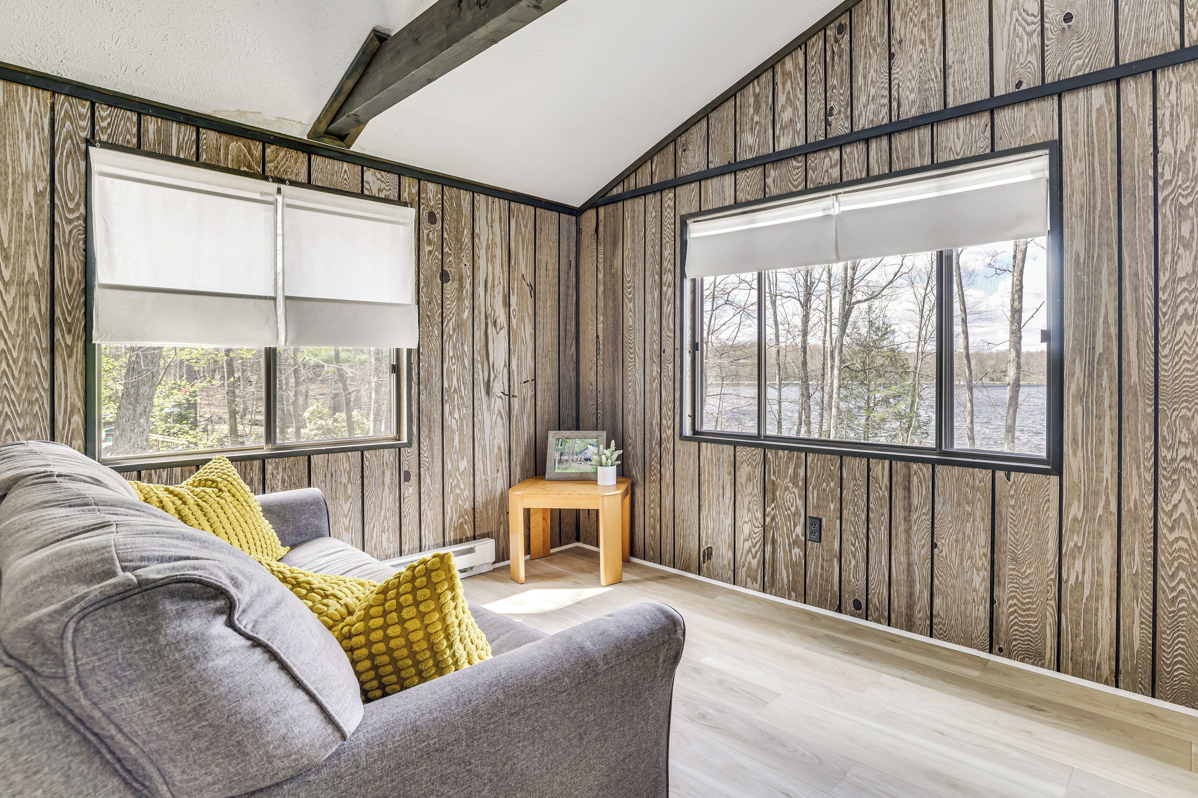 Ein Zimmer mit Holzwandverkleidung, einem grauen Sofa mit gelben Kissen, einem kleinen Holztisch mit Bilderrahmen und großen Fenstern mit Jalousien.