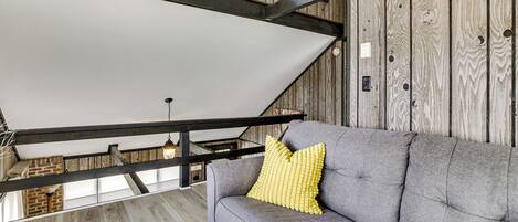 Una habitación estilo loft con vigas de madera a la vista, un sofá gris con un cojín amarillo y un ventilador de techo.