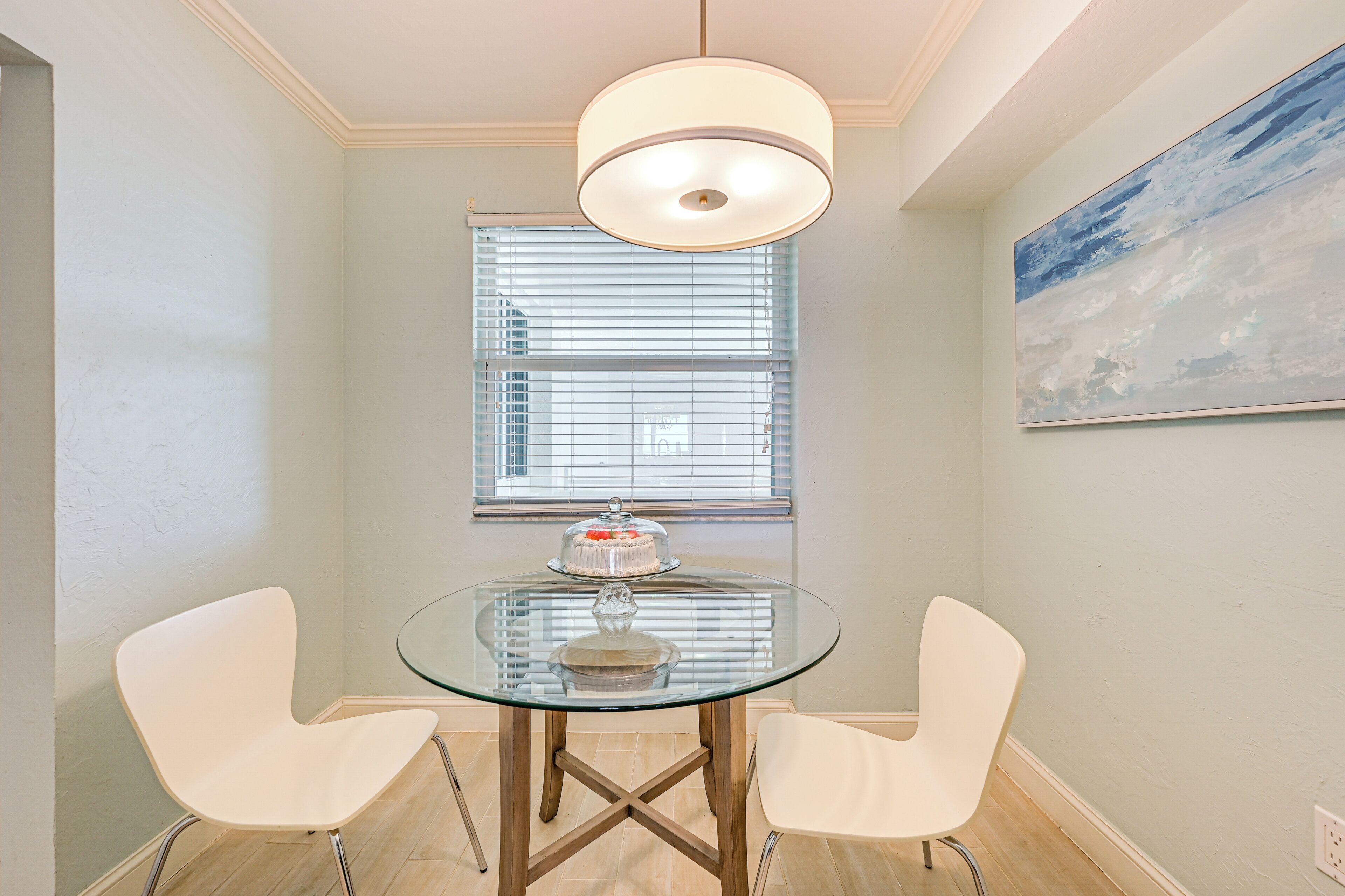 A dining area with a glass table, two white chairs, a ceiling light, a window with blinds, and a painting on the wall.