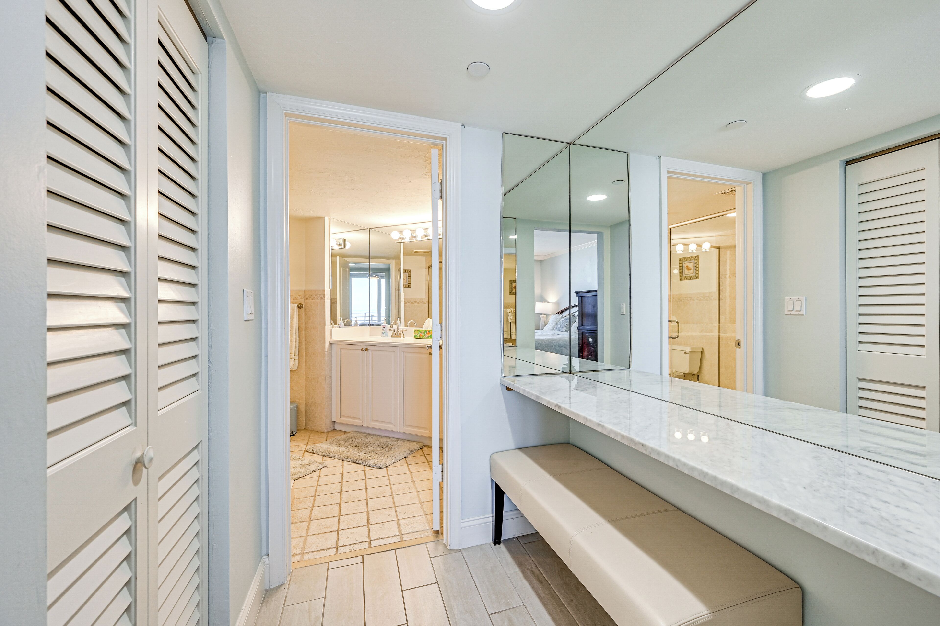 A bathroom with a vanity, mirror, bench, and a view into the adjacent bedroom.