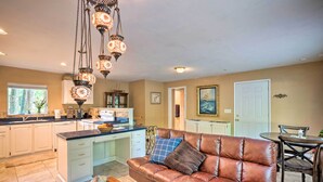 A spacious living area with a brown leather sofa, a wooden coffee table, and a dining table with chairs. A kitchen with white cabinets and a large island is visible in the background. - Quaint Riverside Reprieve w/ Kayaks + Floats! (Brookings)