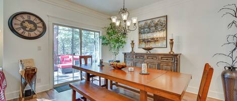 Un coin repas avec une table en bois, des bancs et un lustre. Il y a une horloge au mur et un tableau au-dessus des placards.